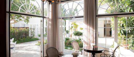 A sunlit room with large windows, wicker chairs, a round table with a vase, and a chandelier.