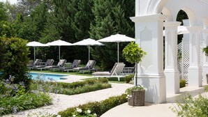 Un espace piscine doté de chaises longues et de parasols, entouré de verdure et agrémenté d’un gazebo blanc.