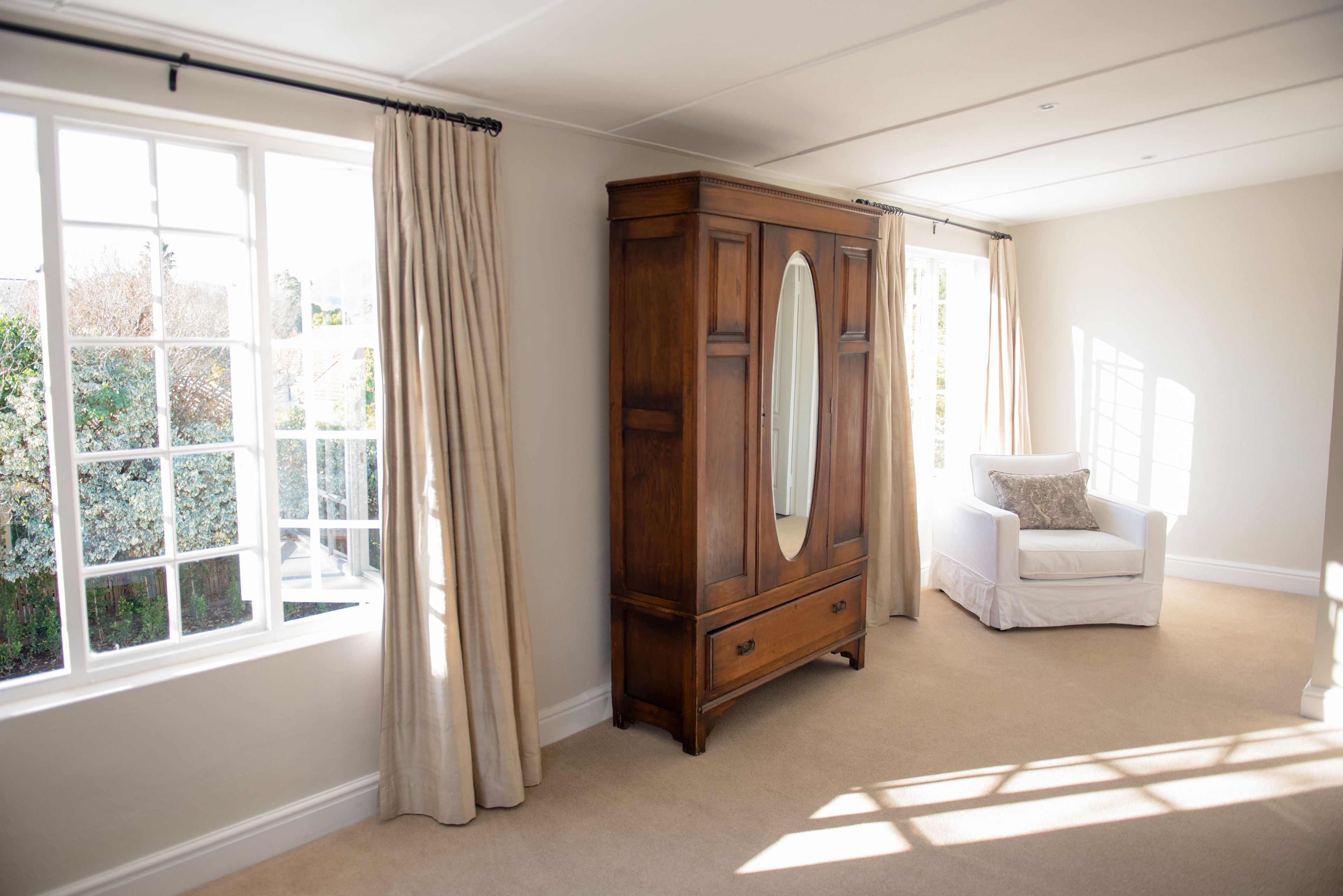 A room with a large window, a wooden wardrobe, a white armchair, and a carpeted floor.