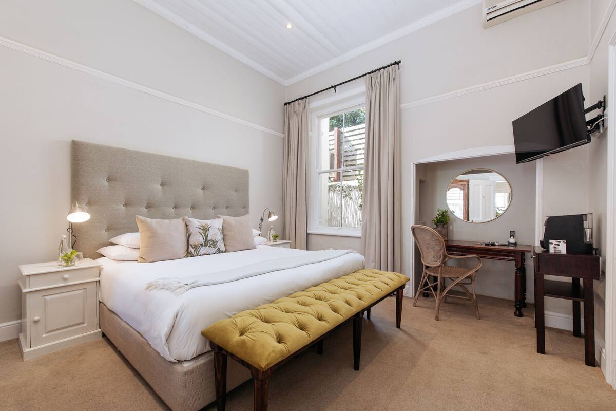 Un dormitorio con una cama grande, una mesita de noche con lámpara, un escritorio con silla, un televisor y una ventana con cortinas.