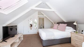 A bedroom with a sloped ceiling, a bed with pillows, a bedside table with a lamp, a TV on a wooden stand, and a wardrobe.