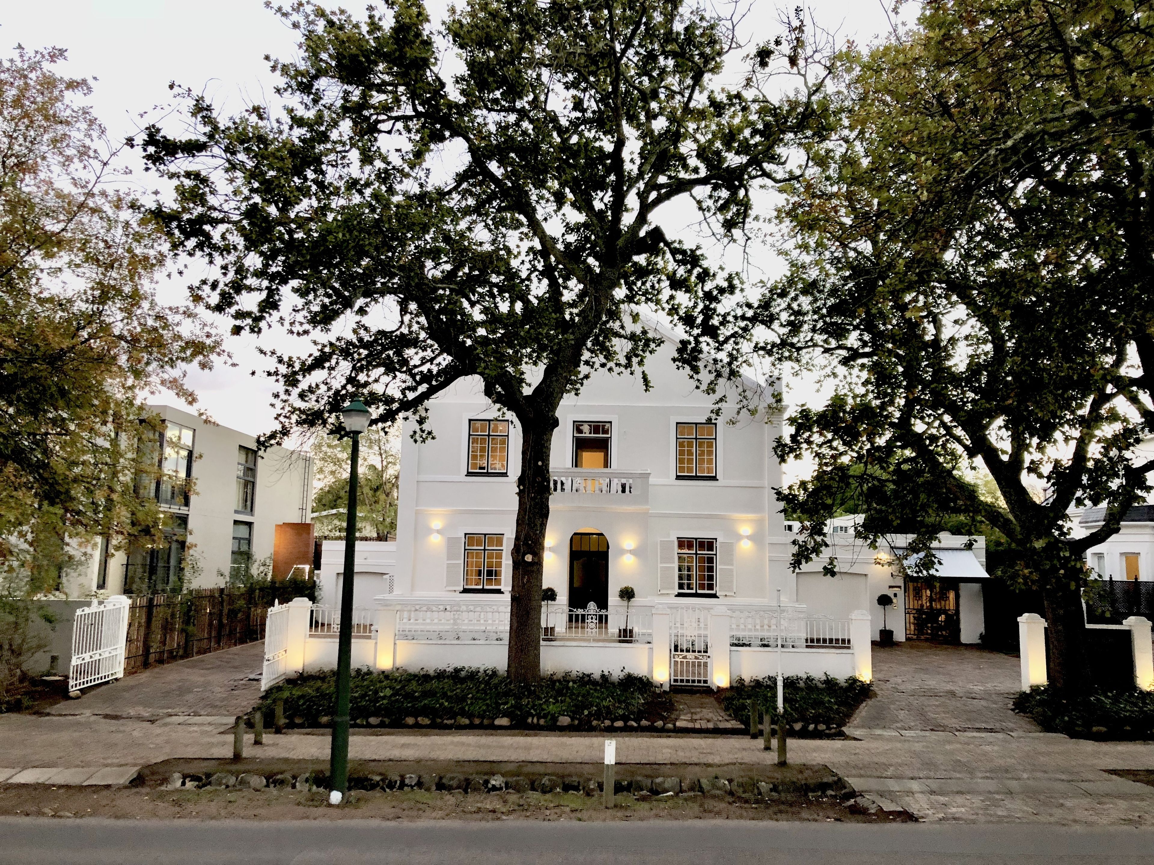 A white building with a large tree in front, a gated entrance, and a paved driveway.