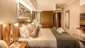 A modern hotel room with a neatly made bed, a bedside table with a lamp, a mirror, and a framed artwork on the wall.