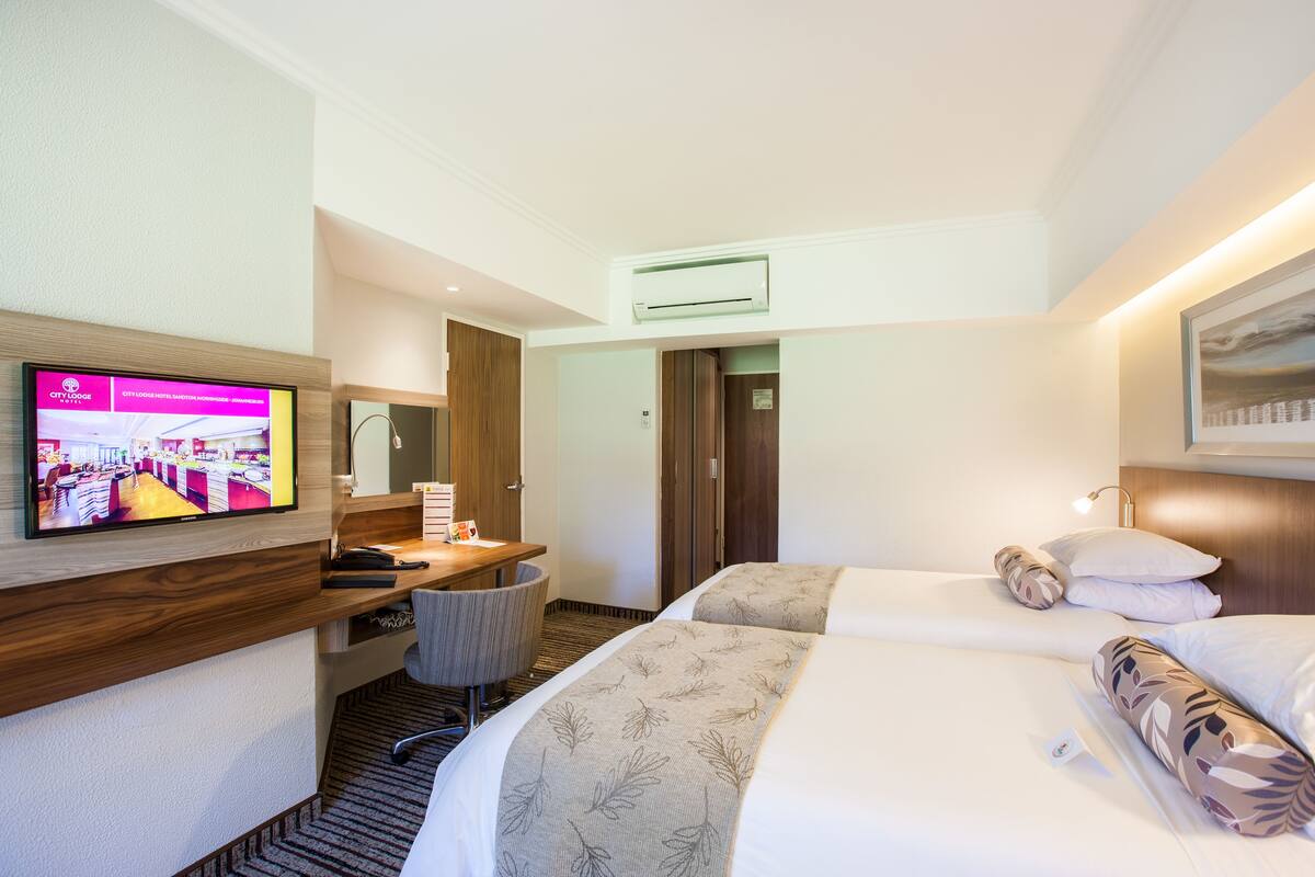 A hotel room with a TV on the wall, a desk with a chair, two beds with floral-patterned bedding, and a wooden headboard.