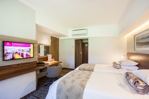 A hotel room with a TV on the wall, a desk with a chair, two beds with floral-patterned bedding, and a wooden headboard.