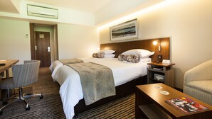 Una habitación de hotel con cama, escritorio, silla, una mesita y una estantería en la pared.