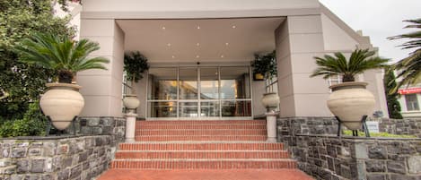 A building with a modern architectural style, featuring a set of stairs leading to a glass entrance, stone walls, and large planters with greenery.