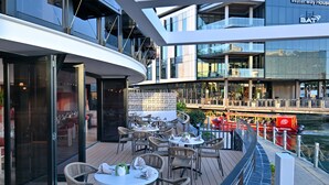 Un espace de restauration extérieur moderne, avec des chaises et des tables en osier, offrant une vue sur une rivière et un pont.