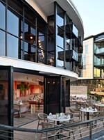An outdoor dining area with round tables and wicker chairs, a bar area with stools, and a modern building with large windows and a balcony.