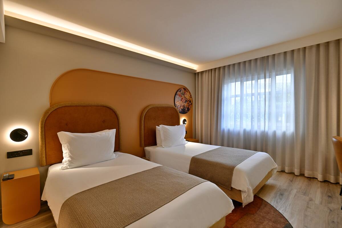 Ein Hotelzimmer mit zwei Betten, einer runden Wanduhr, einem Nachttisch und einem Fenster mit Vorhängen.