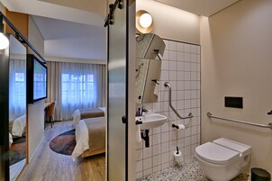 A hotel room with a bed, a desk, and a TV. It also shows a bathroom with a sink, toilet, and shower.