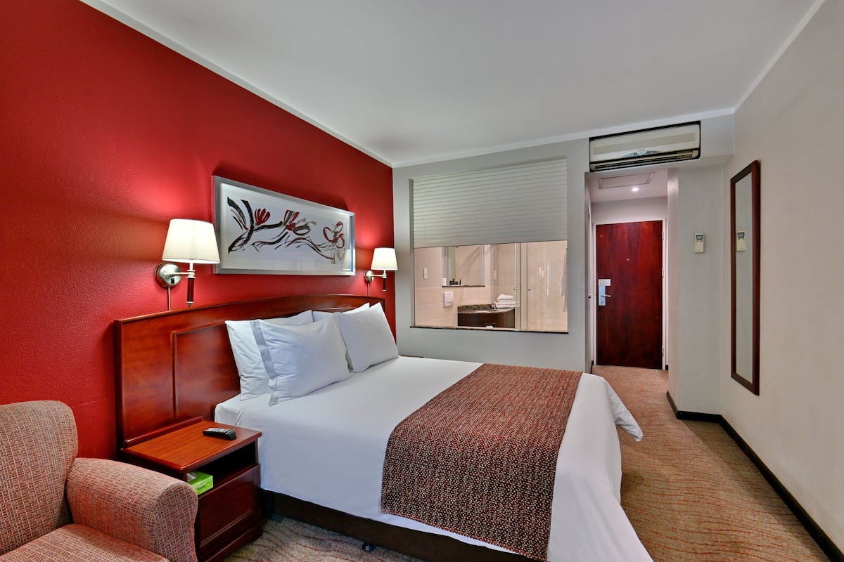 Habitación de hotel con una cama grande, dos lámparas de noche, una mesita de noche, una silla, una pared de acento roja, un cuadro, un espejo y un baño visible a través de una puerta abierta.