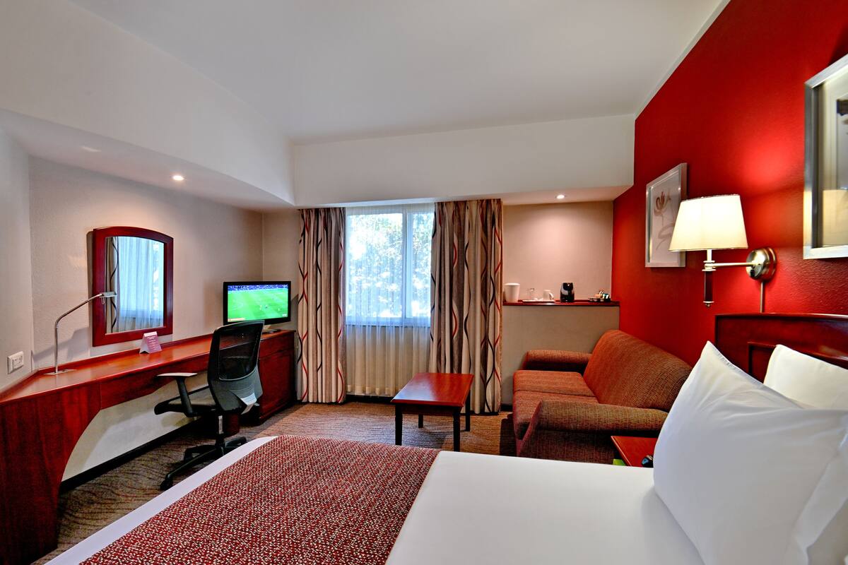 Une chambre d’hôtel équipée d’un lit, d’un bureau, d’une chaise, d’une télévision et d’un mur d’accent de couleur rouge.