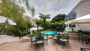 Un patio al aire libre con suelo de ladrillo, mobiliario exterior con mesas y sillas, una piscina y abundante vegetación circundante.
