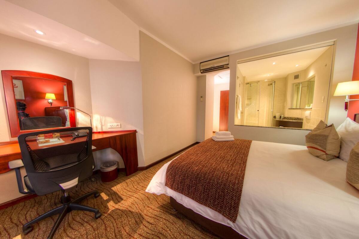 Une chambre d’hôtel avec un lit, un bureau, une chaise, un miroir et une salle de bain visible à travers une porte ouverte.