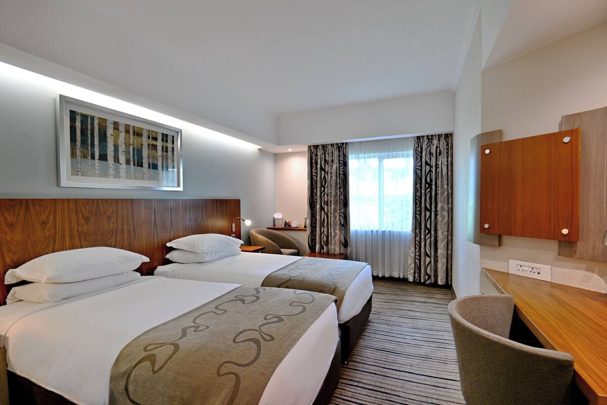 Ein Hotelzimmer mit zwei Betten, einem Schreibtisch, einem Stuhl, einem Fernseher und einem Fenster mit Vorhängen.