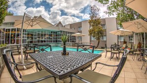 An outdoor pool area with a table and chairs, surrounded by umbrellas and a building with large windows.