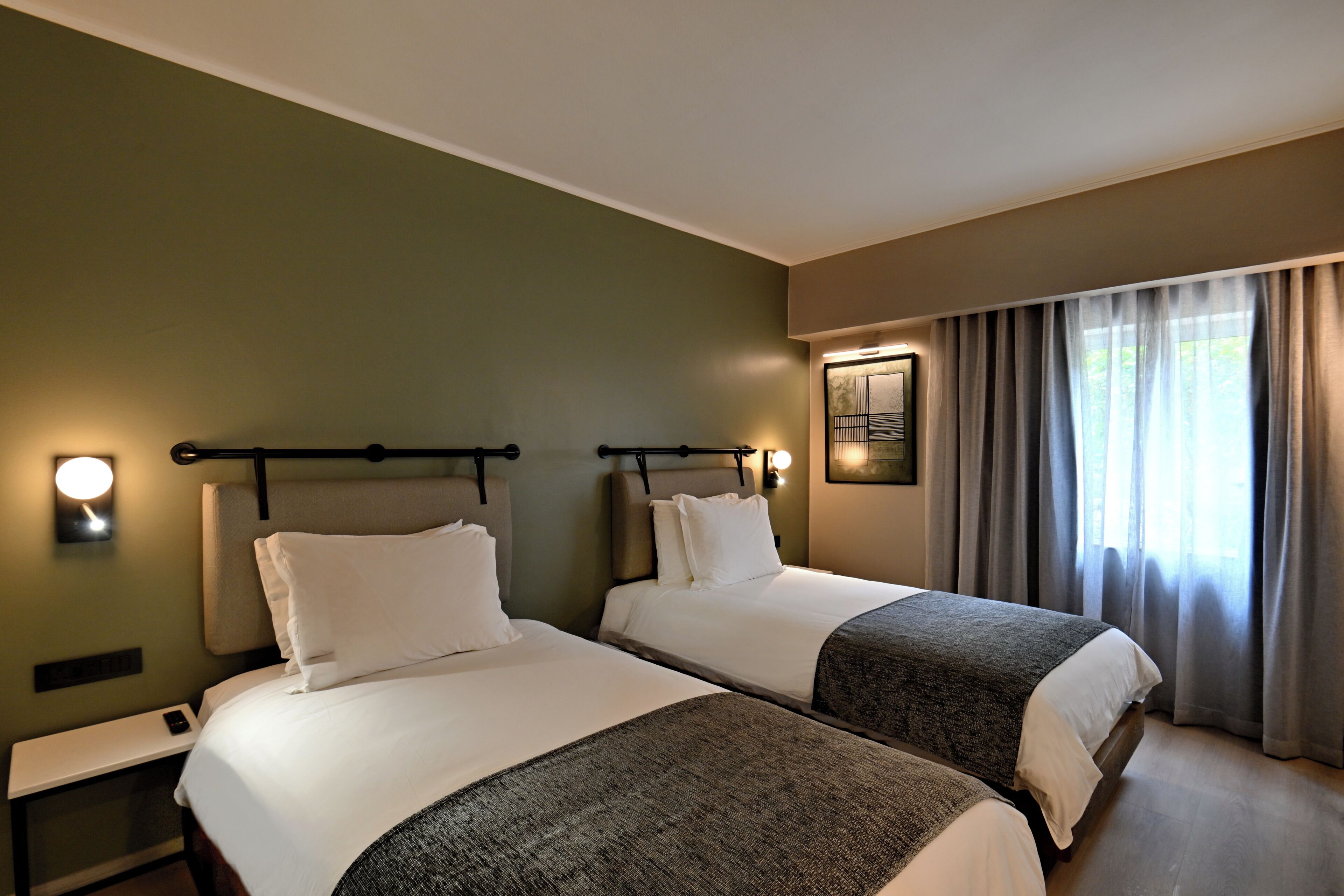 A hotel room with two beds, a nightstand, a wall-mounted lamp, and a window with curtains.