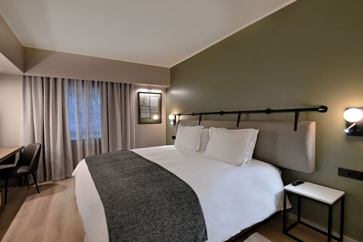 Et hotelværelse med seng, skrivebord, stol, natbord og et vindue med gardiner.