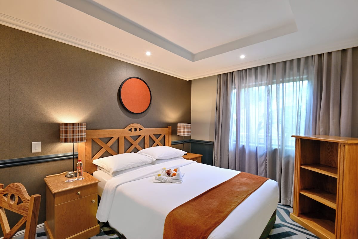 A hotel room with a bed, a wooden nightstand, a chair, a round wall decoration, and a window with curtains.