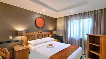 Habitación de hotel con cama, mesita de noche de madera, una silla, un adorno circular en la pared y una ventana con cortinas.