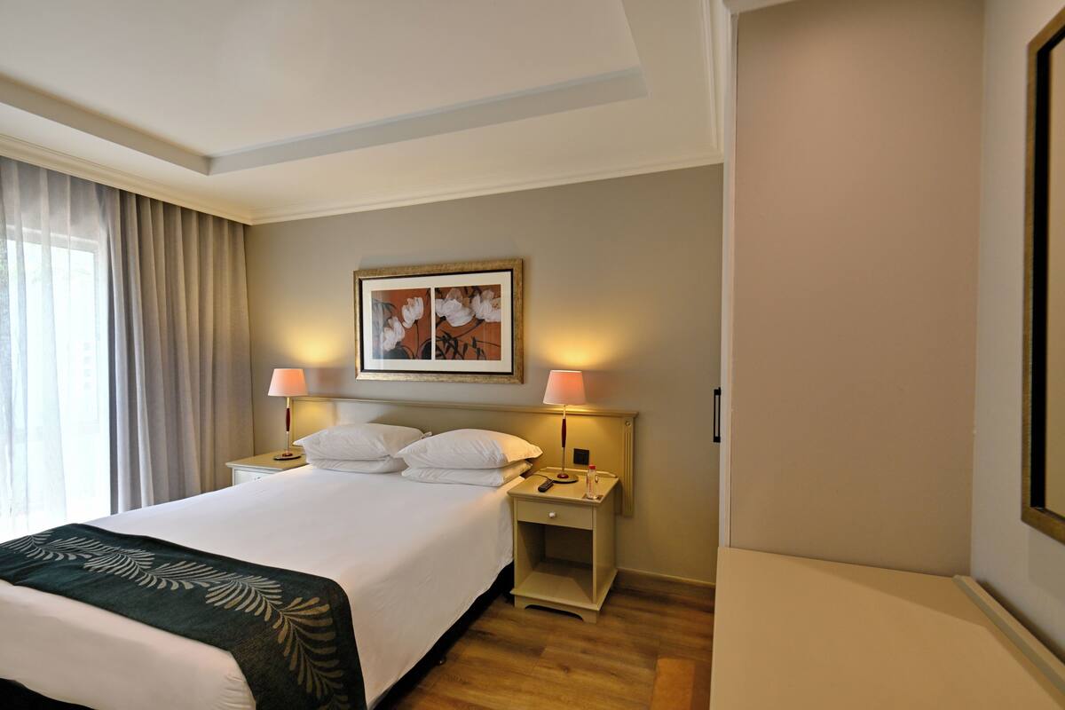 Een hotelkamer met een groot bed, twee nachtkastjes, een lampje en een kunstwerk boven het bed, plus een raam met gordijnen.