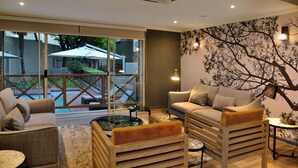 A modern living room with a sofa, armchairs, and a coffee table. There is a glass door leading to an outdoor area with umbrellas. - Courtyard Hotel Sandton (Sandton)