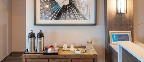 A reception desk with a framed cityscape artwork, a coffee station with thermos and cups, and a sign indicating a public area.