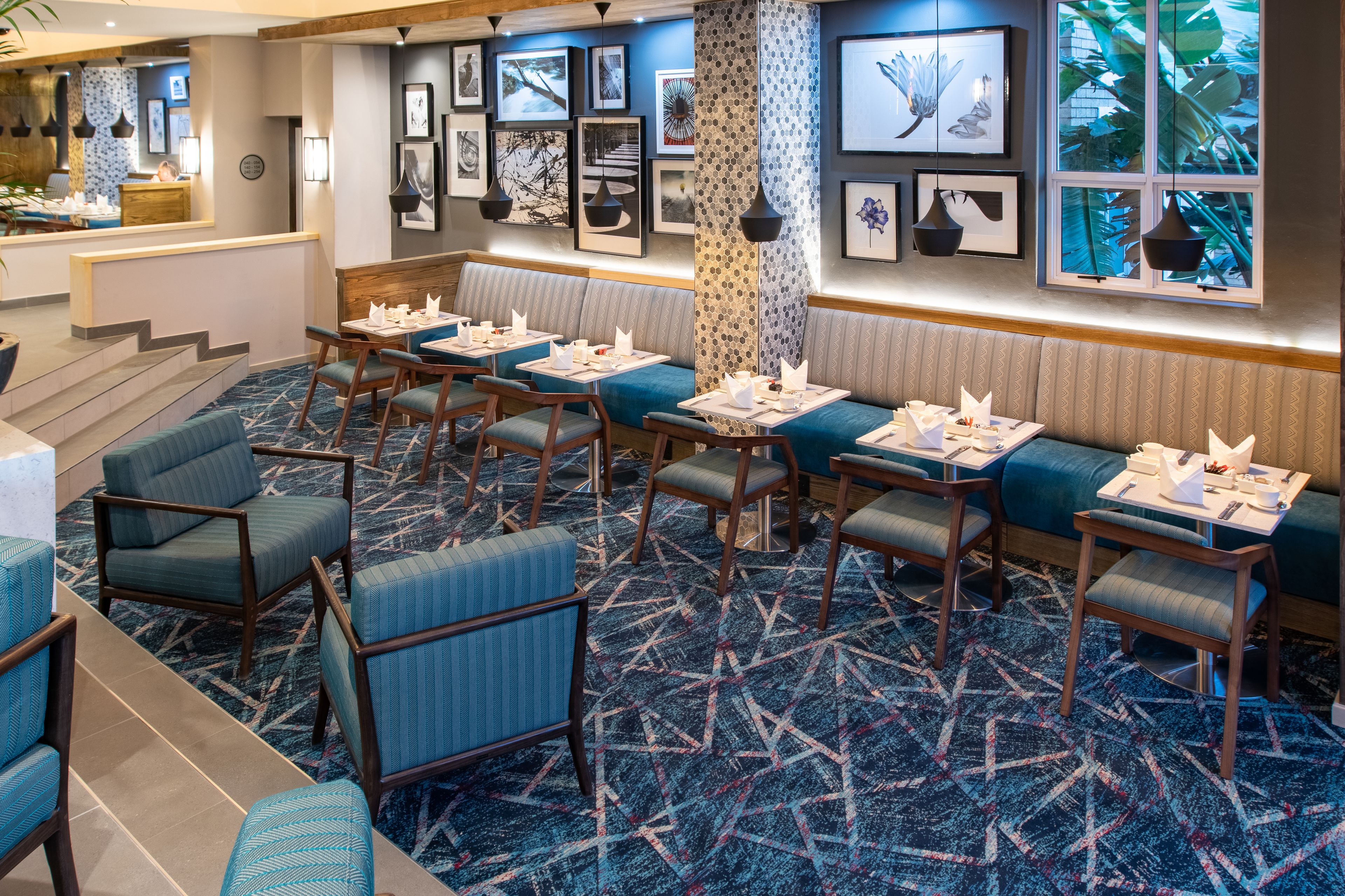 Un hall de hotel con una zona de estar que incluye sillones tapizados en azul y mesas, una alfombra estampada y cuadros enmarcados en las paredes.