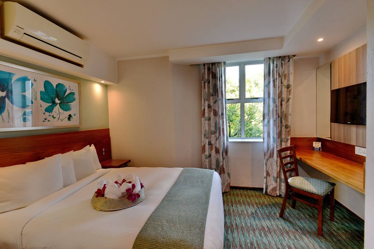 Habitación de hotel con cama, escritorio, silla, aire acondicionado, ventana con cortinas y televisión de pantalla plana.