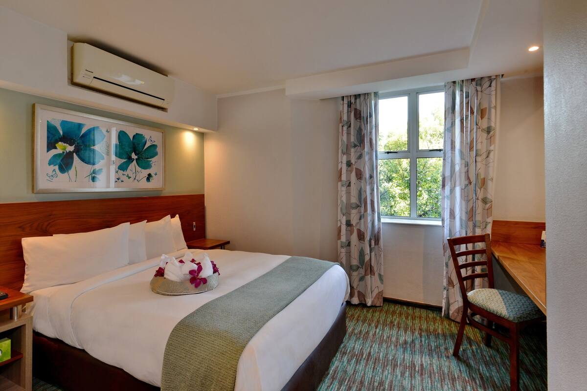Una habitación de hotel con una cama grande, cabecera de madera, un escritorio con silla, ventana con cortinas y aire acondicionado de pared.