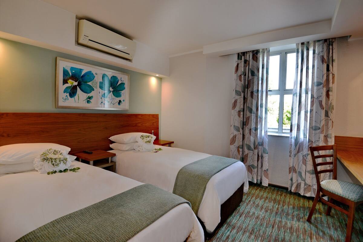 Habitación de hotel con dos camas, cabecera de madera, cuadro floral enmarco, una mesita con un control remoto y una ventana con cortinas estampadas.