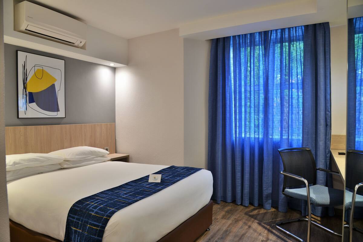 A hotel room with a bed, a desk, a chair, and a large window with blue curtains.