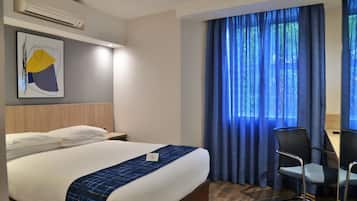 Habitación de hotel con cama, escritorio, silla y un amplio ventanal con cortinas azules.