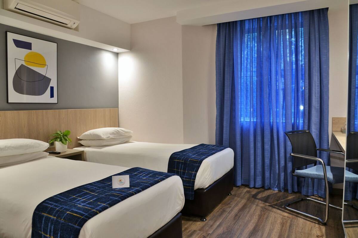 A hotel room with two beds, a desk with a chair, a potted plant, and a large window with blue curtains.