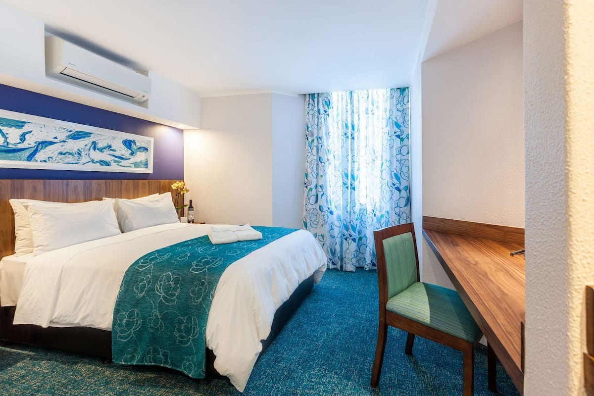 A hotel room with a bed, a desk, a chair, a window with curtains, and an air conditioning unit.