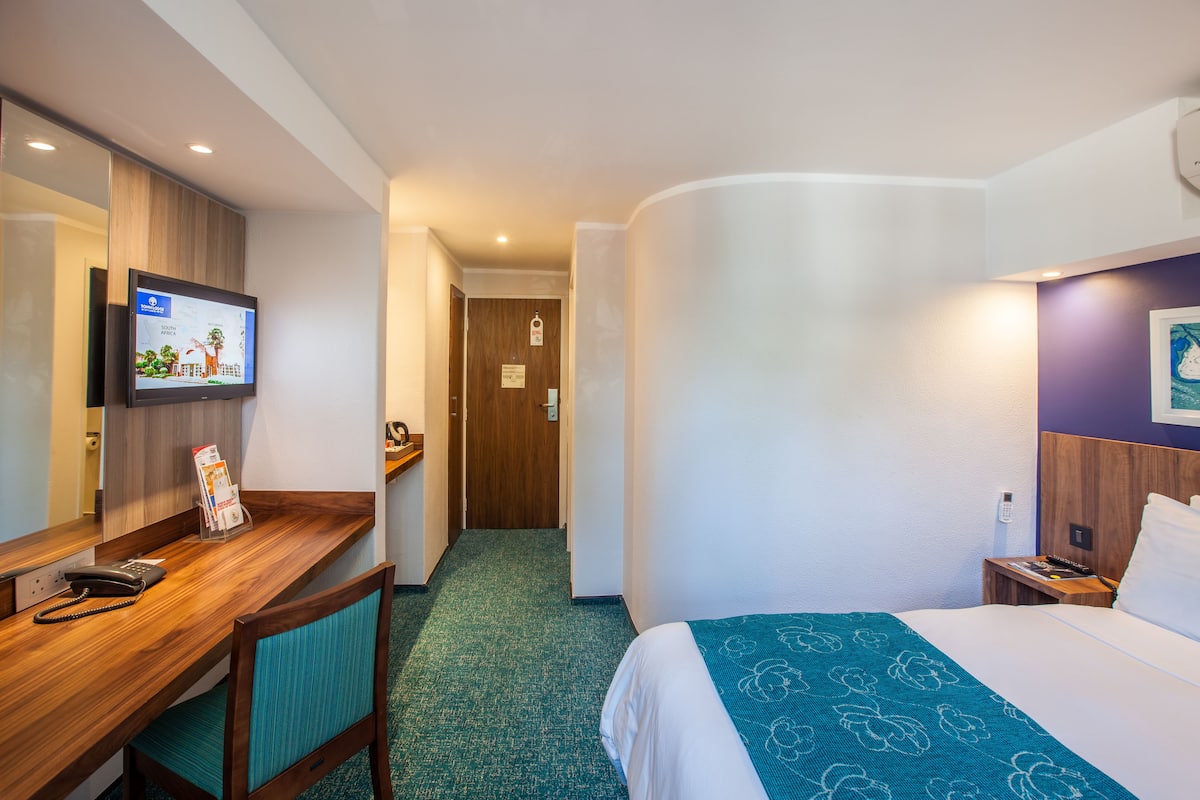 A hotel room with a bed, a desk with a telephone, a television mounted on the wall, and a wooden door leading to the hallway.