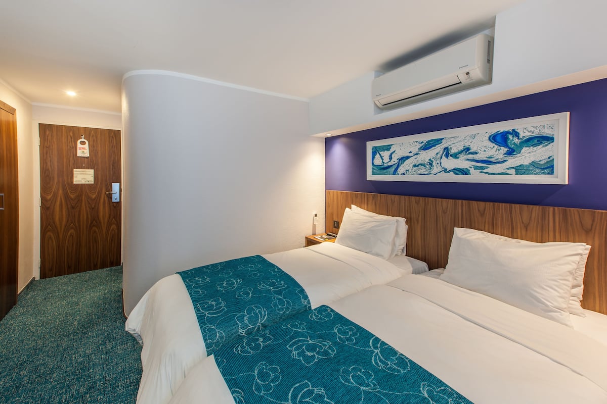 A hotel room with a bed, a wooden headboard, a wall-mounted air conditioner, and a framed artwork above the bed.