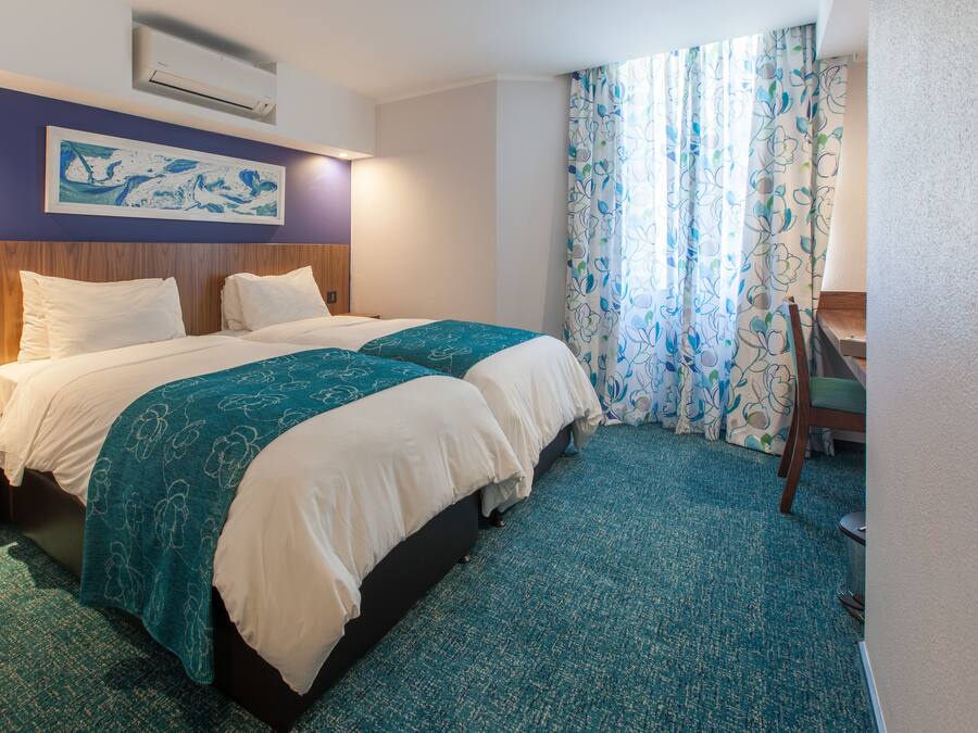 A hotel room with a double bed, a desk, a chair, and a large window with curtains.