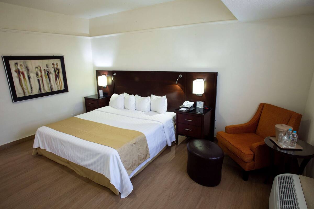 A hotel room with a large bed, a chair, a table with a lamp, and a painting on the wall.