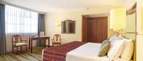 A hotel room with a bed, a desk, two chairs, a mirror, and a lamp.