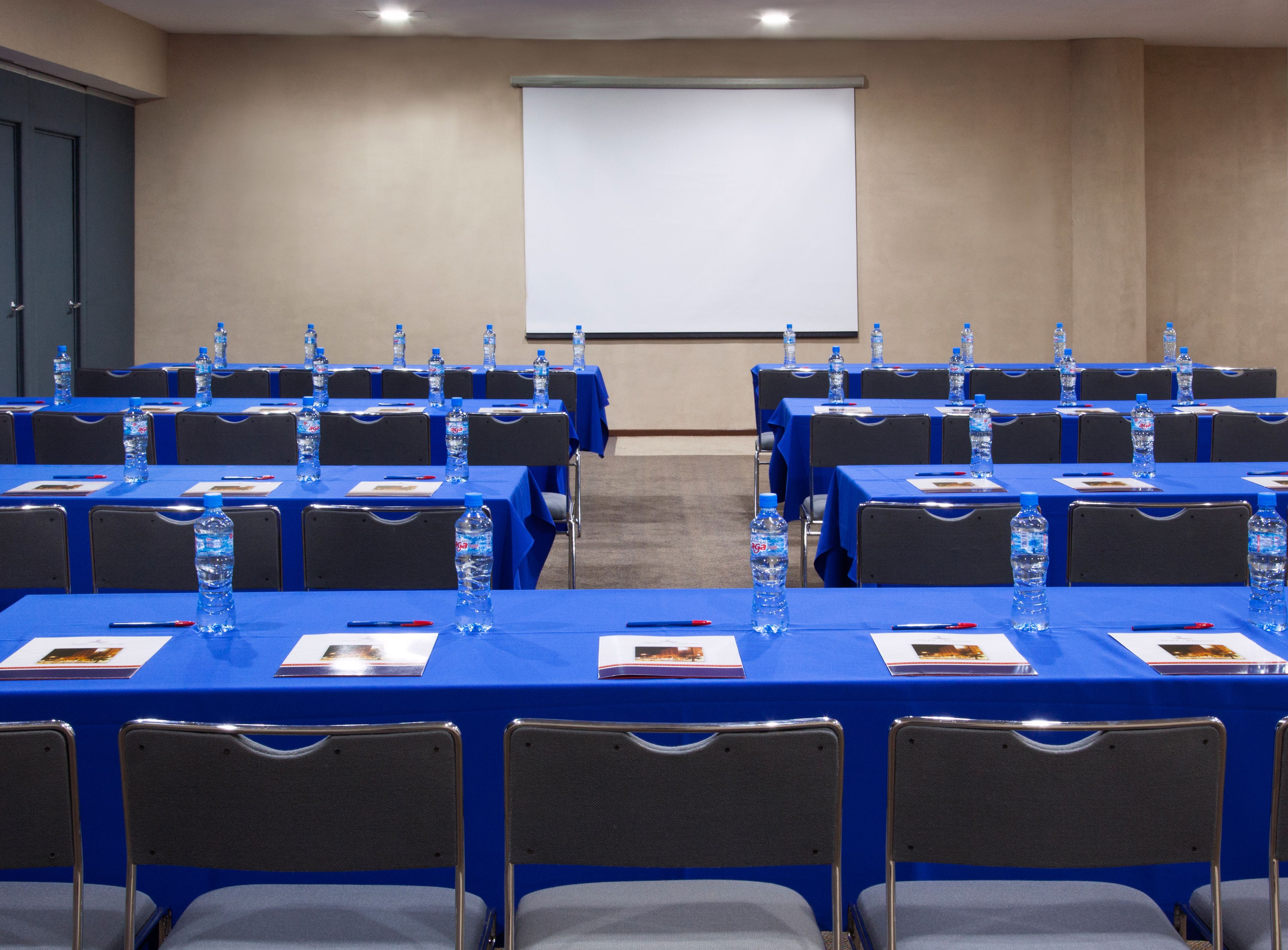 A conference room with rows of tables, each with water bottles and nameplates, and a projector screen on the wall.