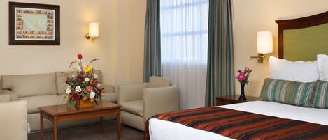 Una habitación de hotel con una cama, dos sillones, una mesita de café, una lámpara, un cuadro en la pared y una ventana con cortinas.