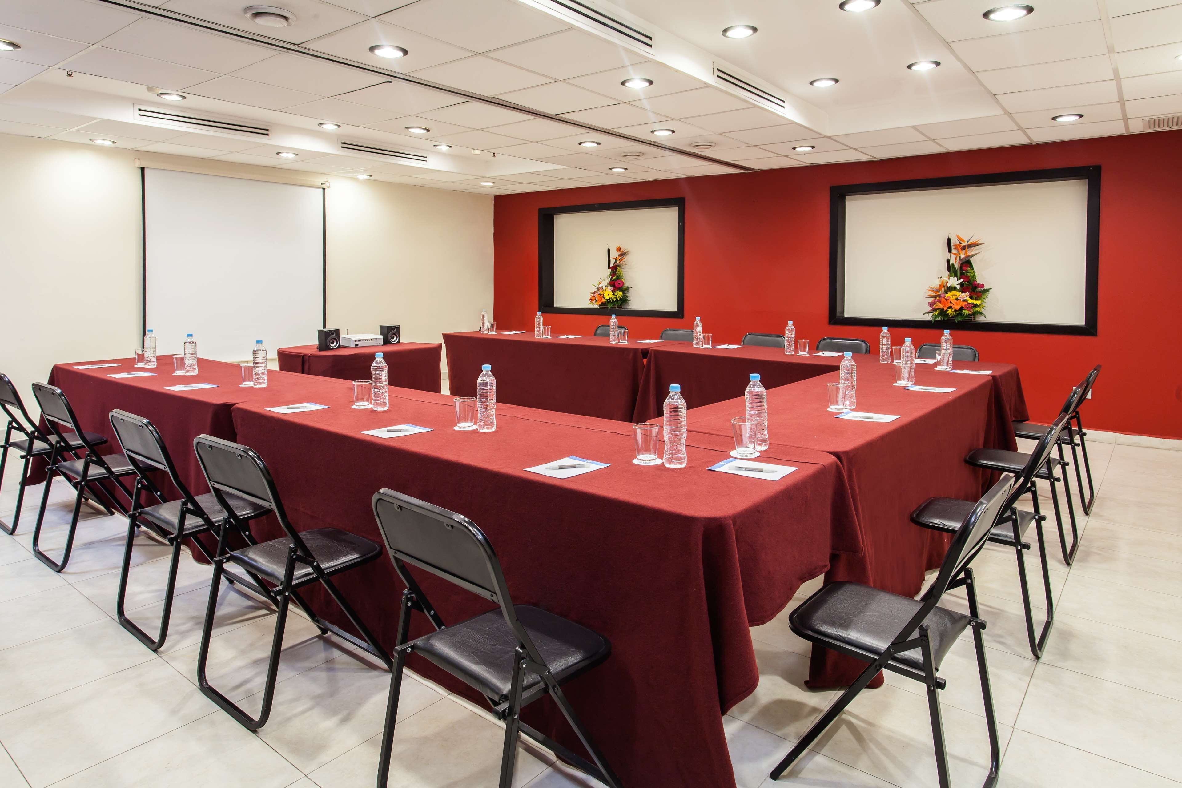 Una sala conferenze con tovaglie rosse, sedie pieghevoli, bottiglie d'acqua e un proiettore.