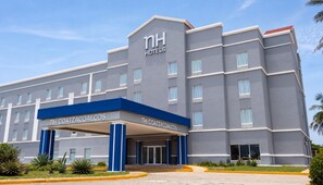 Un edificio de hotel con diseño moderno, que presenta una entrada destacada con un toldo azul y el nombre 'NH COATZACOALCOS' a la vista.