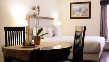 Habitación de hotel con una cama, dos sillas, una mesa con fruta y una planta, y un cuadro en la pared.