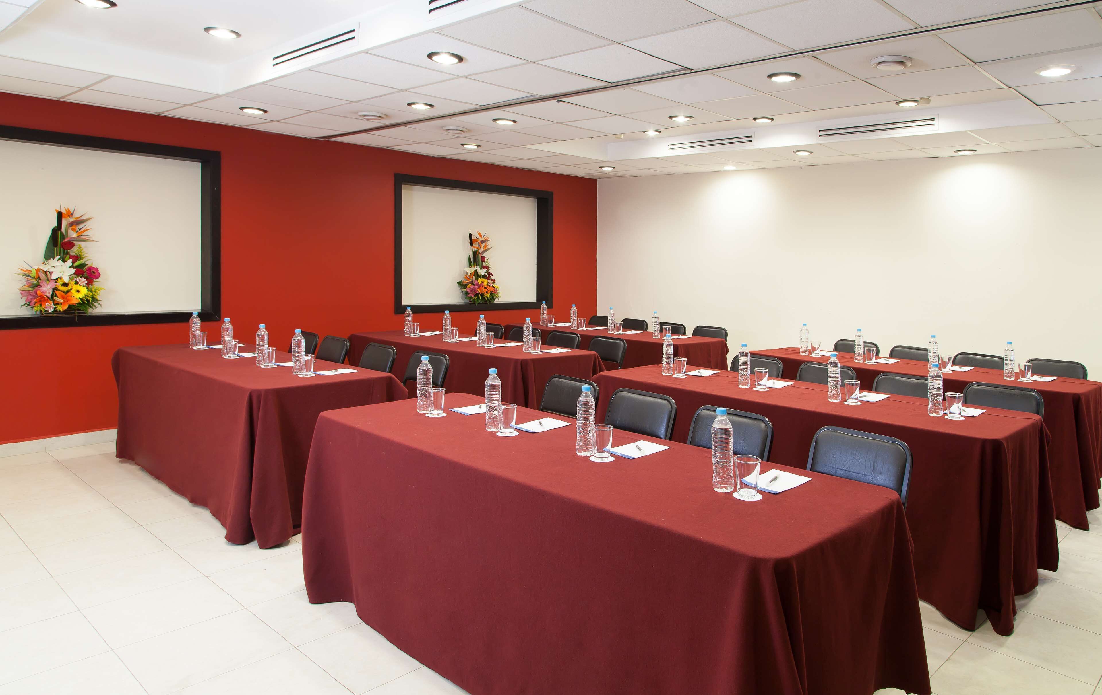 A conference room with multiple tables covered in red tablecloths, equipped with water bottles and glasses, and featuring a projector screen and floral decorations.