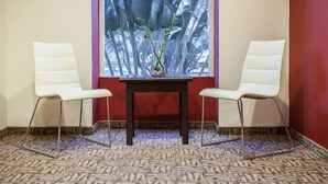 Una habitación con dos sillas blancas, una mesita y un ventanal con vistas a plantas altas.