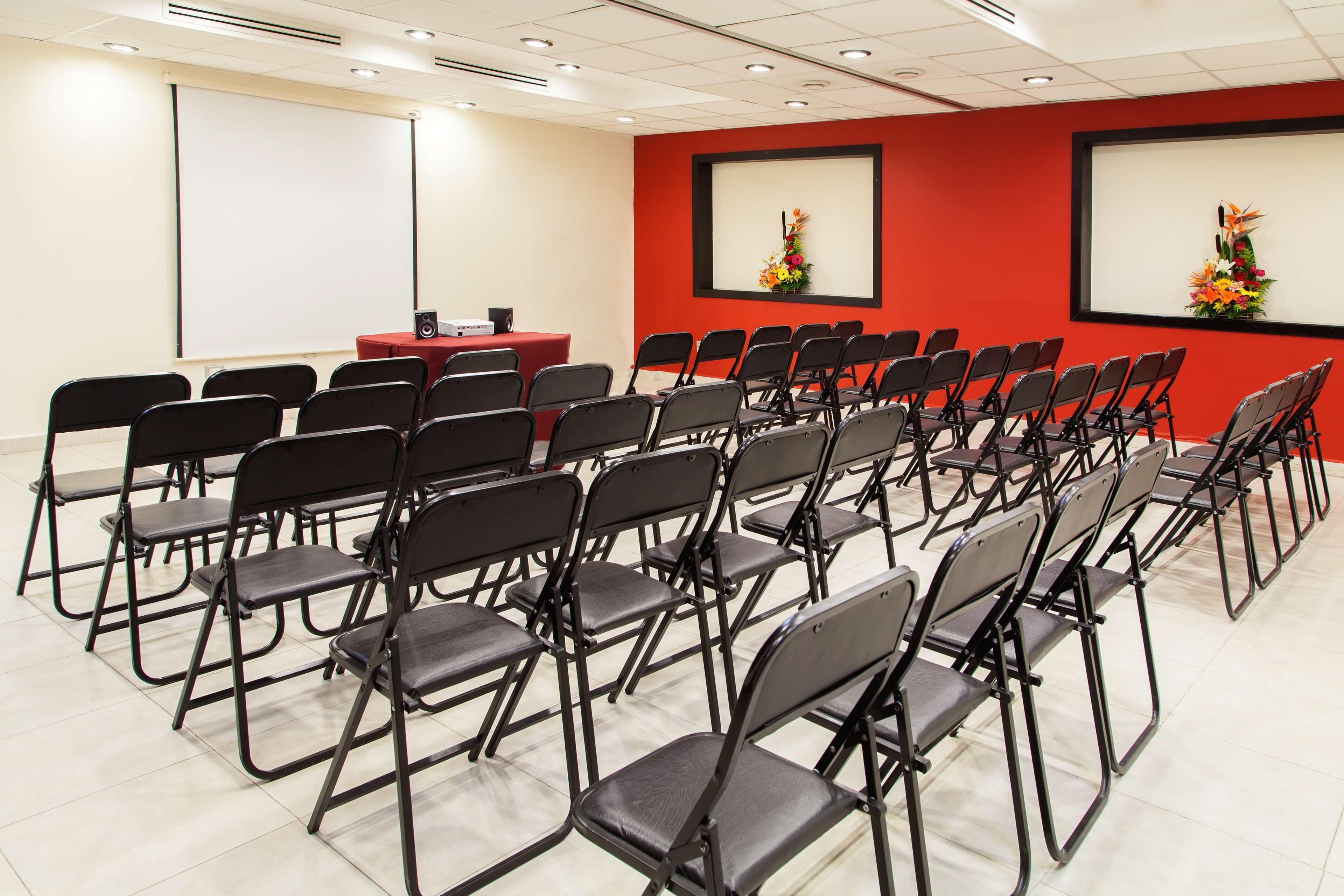 A conference room with rows of foldable chairs, a projector screen, and a red accent wall with flower arrangements.
