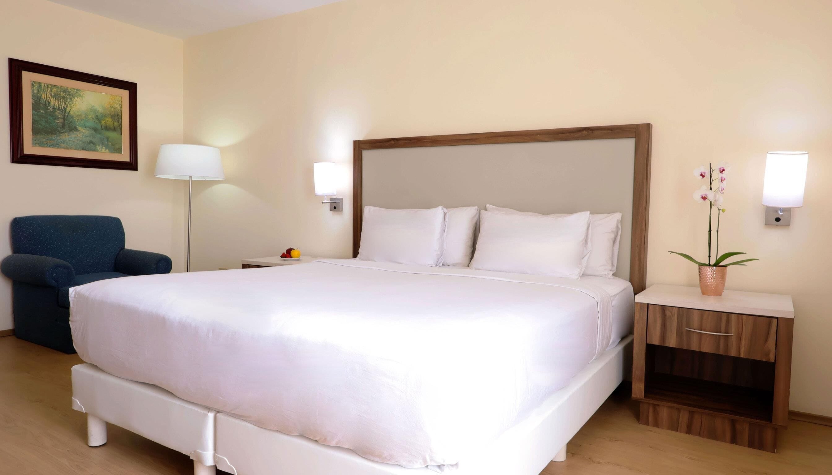 Una habitación de hotel con una cama grande, una silla, una mesita de noche con una lámpara y una luz en la pared.